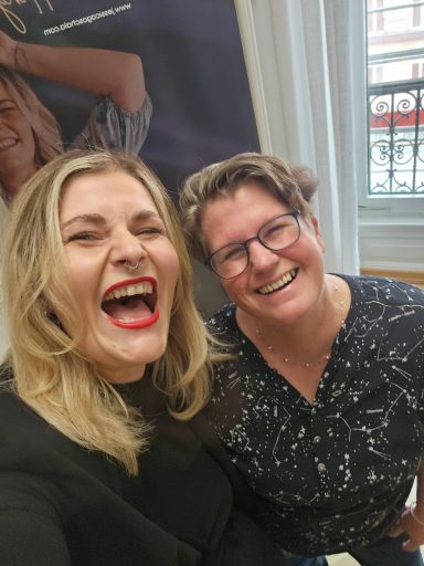 Zwei Frauen lächeln und lachen, stehen nebeneinander vor einer hellen Wand.