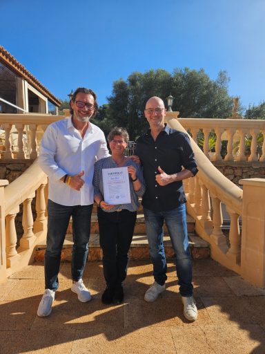 Drei Personen posieren lächelnd auf einer Treppe im Freien, halten ein Zertifikat.