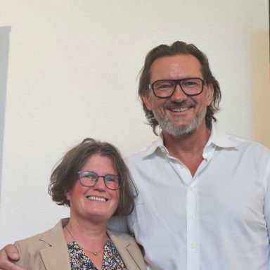 Zwei fröhliche Personen stehen nebeneinander, lächeln und tragen modische Kleidung.